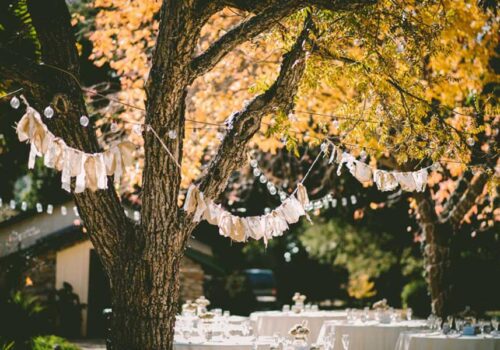 A rustic destination wedding setup with tables and chairs arranged under trees adorned with string lights and garlands. The leaves on the trees have turned golden, creating an inviting and warm autumn atmosphere. Image