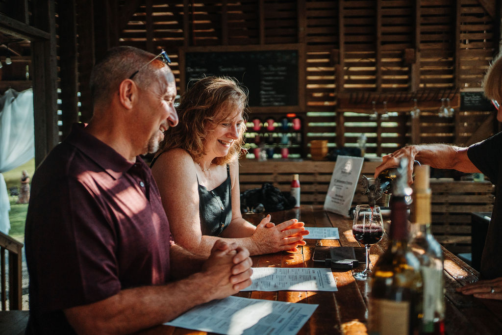 A man and woman smile while sitting at a wooden bar in a rustic setting, filled with natural light. The woman in a black top has her hands clasped, and the man in a burgundy shirt has his hands resting on the bar. A person is pouring wine into a glass in front of them.