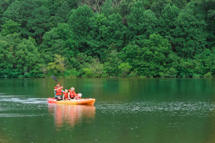 A group of four people, wearing life jackets, are paddling a canoe on a calm lake surrounded by lush, green trees. They appear to be enjoying the tranquil environment of the forested area. The water reflects the greenery, adding to the serene ambiance.