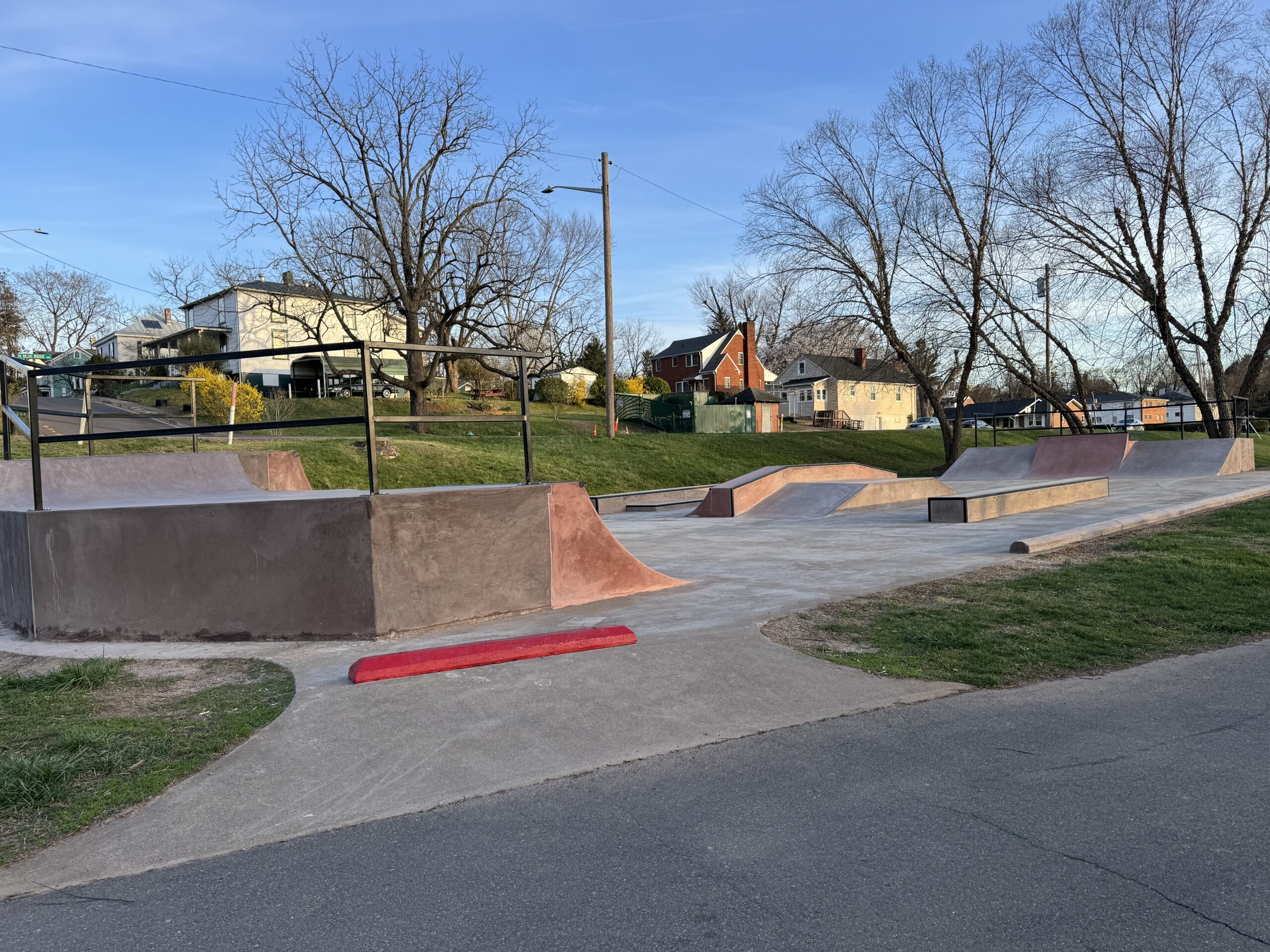 A skate park with various ramps and rails is situated near a residential area with houses and leafless trees. The park features concrete surfaces, a red curb at the entrance, and grassy patches around it, under a blue sky with light clouds.