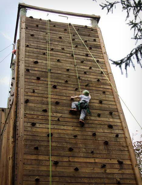 A person wearing a harness and helmet climbs a tall wooden rock climbing wall. The wall is equipped with numerous small handholds and footholds, and several green safety ropes hang down the front. Trees and sky are visible in the background.