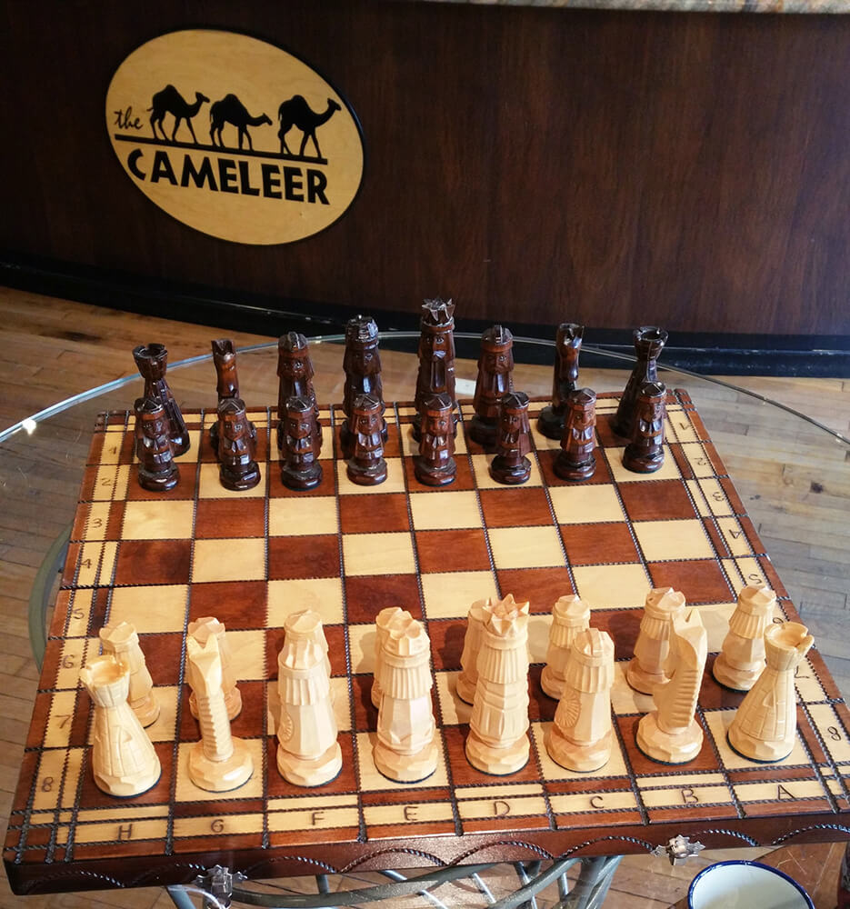 A uniquely designed chess set with detailed dark brown and light wooden pieces on a wooden board with labeled squares is placed on a glass table. In the background, there is a wall featuring 