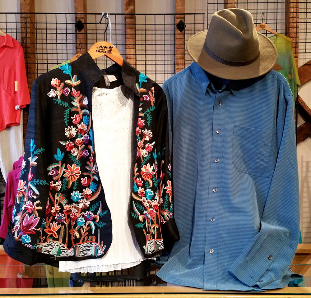 Two shirts on display: a black shirt with colorful floral embroidery and a white blouse underneath on the left, and a blue button-up long-sleeve shirt with a gray fedora on the right. The background features a clothing rack with various hanging garments.