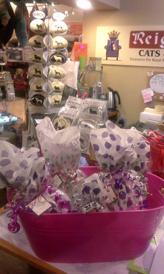 A store display features a bright pink basket filled with several gift-wrapped items in white paper with colorful polka dots. The background includes various cat-themed merchandise and a sign that reads, 