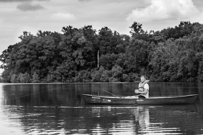 A person sits in a canoe on a calm lake, fishing. The scene is surrounded by dense, leafy trees on the bank, and the sky is partly cloudy. The photograph is in black and white.