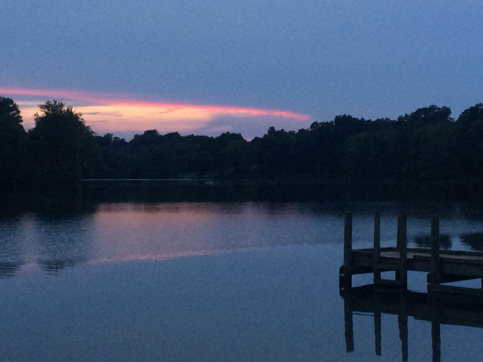 A serene lake at dawn or dusk with calm water reflecting the sky's colors. The horizon features a strip of orange-pink light, transitioning into a mostly cloudy blue sky. Silhouetted trees line the distant shore, and a wooden dock juts into the water on the right.