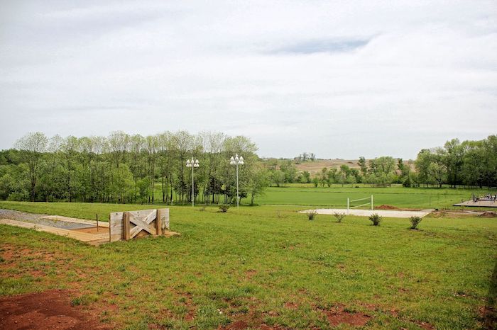 A serene grassy landscape with some small trees in the distance and a few sparsely placed structures, including a wooden pathway and light poles, under a partly cloudy sky. Some areas appear to be under construction or development.