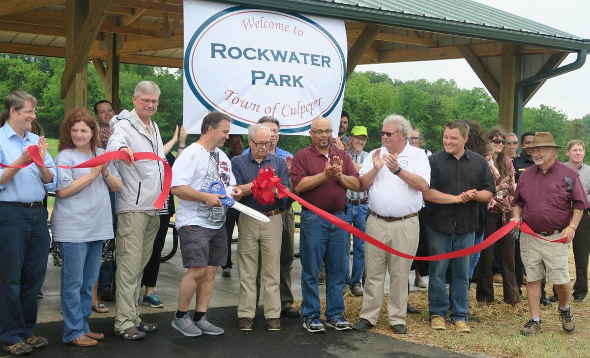 A group of people stands together at the ribbon-cutting ceremony for Rockwater Park in the Town of Culpeper. Participants hold a red ribbon and oversized scissors under a covered pavilion with a large sign reading