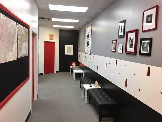 A hallway with white walls, black trim, and red accents. The left wall displays a large map, while the right wall features various framed artworks. Along the right side, there are black benches and small tables. An exit door is visible at the back of the hallway.