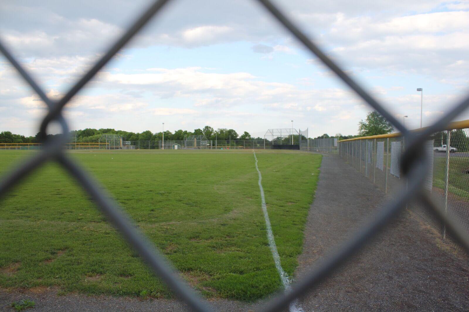 A fenced baseball field with green grass, a dirt path, and metal chain-link fencing is viewed through the diamond-shaped gaps of another fence. The sky above is partly cloudy with scattered clouds, and trees are visible in the background.
