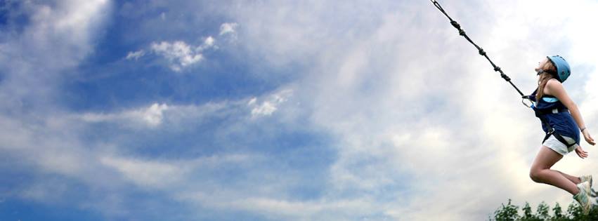 A person wearing a helmet and harness is mid-air, attached to a bungee cord against a backdrop of a partly cloudy sky. Trees are visible at the bottom of the image. The scene conveys a sense of adventure and exhilaration.