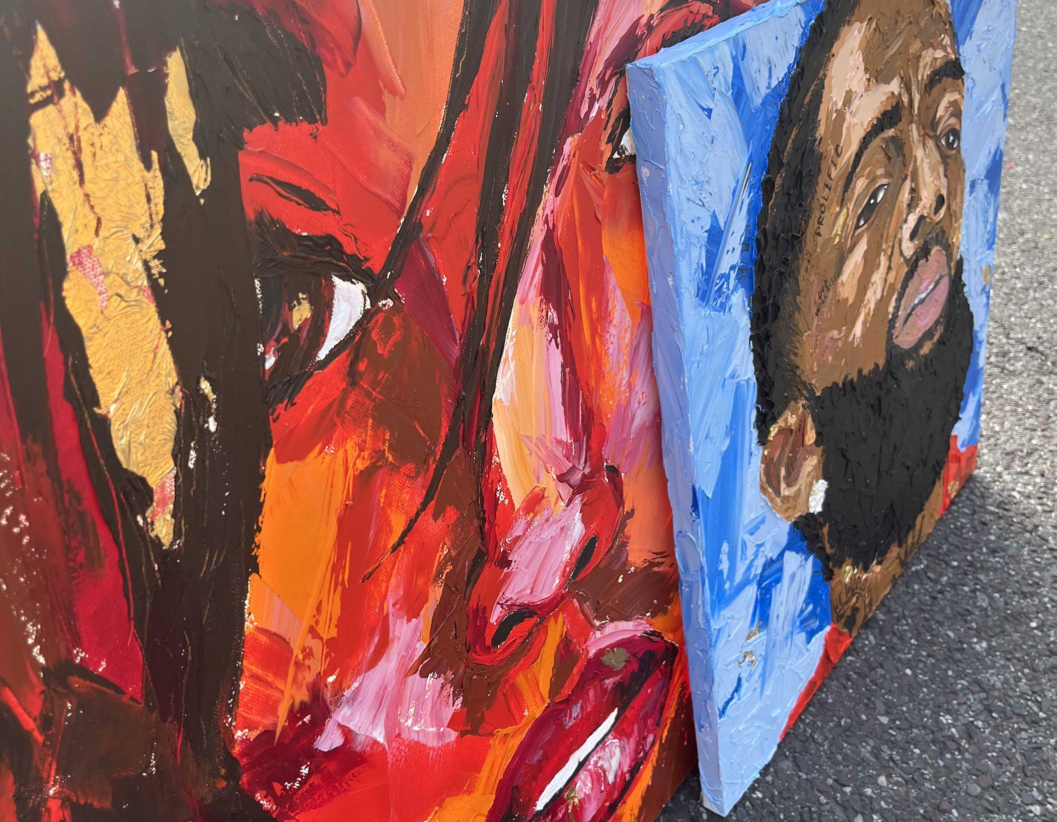 Two vibrant, abstract portrait paintings are leaning against each other. The first features intense red and orange tones, focusing on expressive eyes. The second shows a man with a full beard against a blue background. Both artworks exhibit textured, bold brushstrokes.