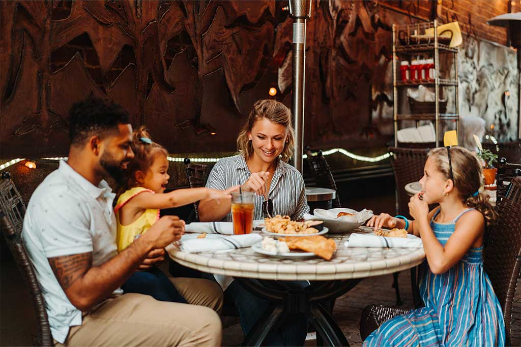 A family of four sits around a round outdoor table at a restaurant, enjoying a meal. The father and daughter on the left are smiling and interacting, while the mother in the center serves food. The daughter on the right, in a blue dress, eats her meal.
