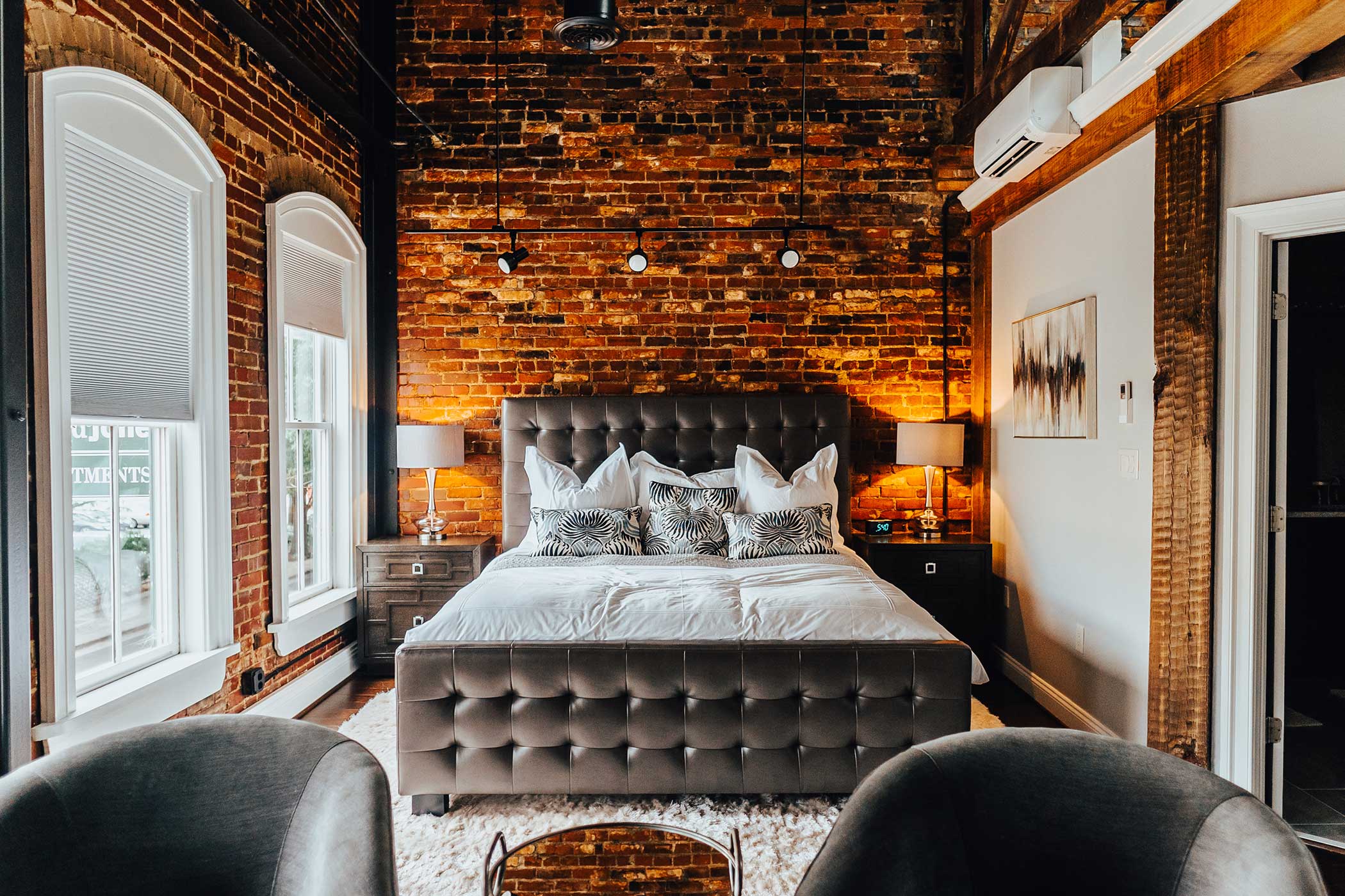 A well-lit bedroom features a large bed with a tufted headboard against a brick accent wall. The bed has multiple pillows and zebra-print cushions. Two bedside tables with lamps flank the bed. Sunlight streams in through two tall windows with white blinds.
