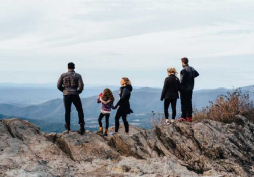 Five people, including three adults and two children, stand on a rocky outcrop overlooking a scenic mountain range. They face away from the camera, taking in the expansive view under a cloudy sky. Some wear jackets and hiking shoes, indicating a outdoor adventure. Image
