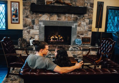 A couple with drinks sits on a brown leather couch, facing a stone fireplace with a warm fire. The rustic room has dark wooden panels, framed windows, and a poster on the wall. They appear cozy and comfortable in the homey setting. Image