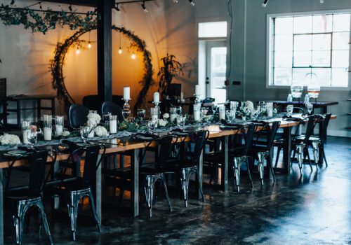 A rustic, elegantly decorated dining area with two long wooden tables set with candles, flowers, glassware, and place settings. Metal chairs provide seating, and an arch adorned with greenery and hanging lights creates a cozy ambiance in the background.