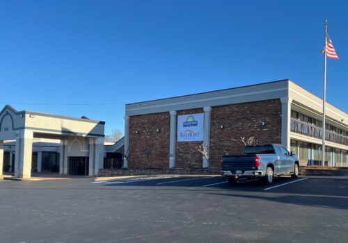 A Baymont hotel building under a clear blue sky. The main entrance is to the left, and an American flag is flying on a pole to the right. A large sign with the Baymont logo hangs on the building. A blue truck is parked in the nearly empty parking lot. Image