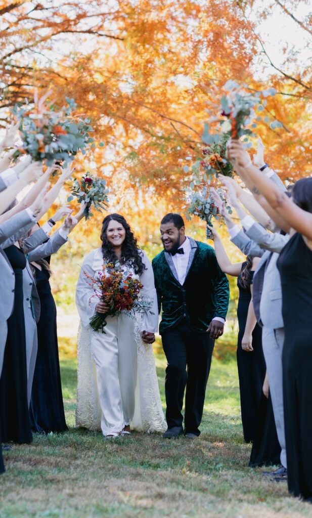 A bride and groom walk hand in hand under an arch formed by raised bouquets held by their wedding party. The bride wears a long white dress, and the groom wears a dark green velvet jacket. The wedding party, dressed in dark outfits, celebrates their union. Autumn trees serve as the backdrop.