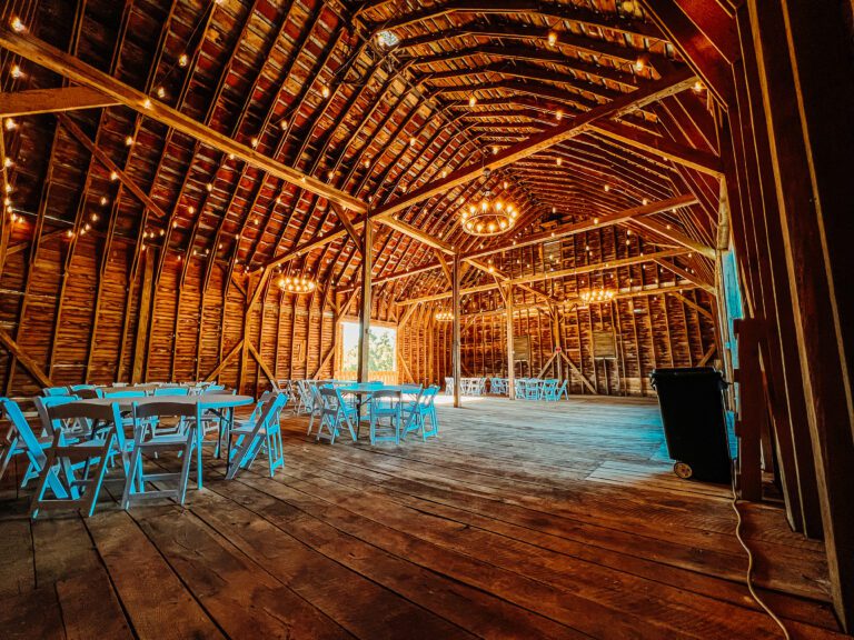 A spacious, rustic barn venue with wooden floors and walls. Bright string lights and chandeliers hang from the vaulted ceiling. Round tables with blue chairs are arranged throughout the space, suggesting it's set up for an event or gathering.