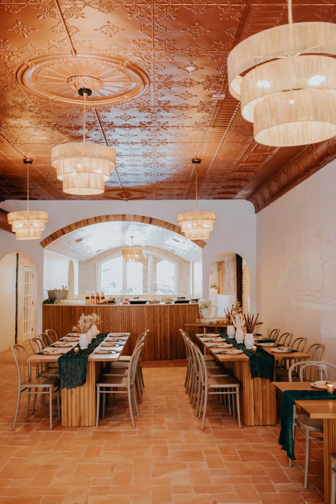 A spacious restaurant interior with warm lighting, featuring a copper-patterned ceiling and multiple pendant lamps. The room has several wooden tables and chairs set for dining, with a bar area in the background. The overall aesthetic is cozy and inviting.