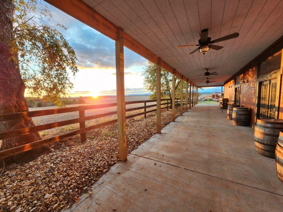 A wide porch with wooden pillars and ceiling fans overlooks a picturesque sunset. The porch has several barrels along the wall and a wooden fence on the left. Trees and a sweeping landscape can be seen in the distance under a partly cloudy sky.