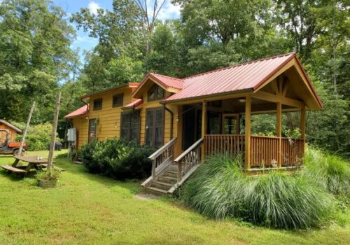 A quaint wooden cabin with a red metal roof, surrounded by tall trees and greenery. The cabin has a covered porch with steps leading up to it. A small wooden structure is seen to the left, and a picnic table is situated on a grassy lawn in the foreground. Image