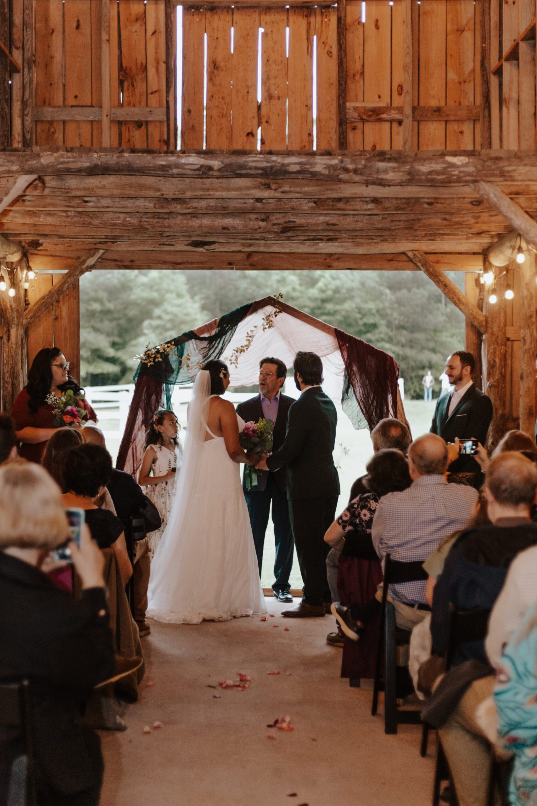 A couple stands facing each other, holding hands, beneath a rustic wooden arch adorned with draped fabric during their wedding ceremony in a charming barn at a premier Rustic Wedding Venue in Virginia. Guests are seated on either side, and the officiant stands in the center, conducting the ceremony.