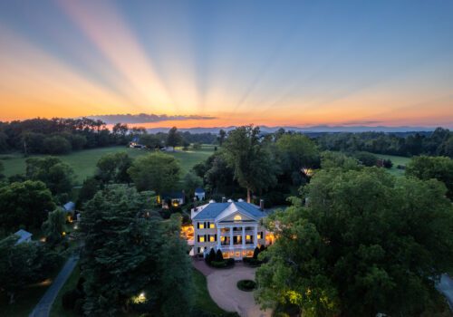 Aerial view of a large, illuminated mansion with columns, surrounded by trees and greenery at dusk. The sky glows with vibrant hues of orange and pink, and sun rays extend through the clouds, enhancing the serene countryside backdrop. Image