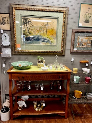 A wooden display table with two shelves holds various glass figurines and collectibles. Above it hangs a framed landscape painting of a road surrounded by trees. Surrounding the painting are smaller framed artworks and shelves containing other decorative items, perfect for antiques and collectables shopping.