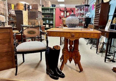 An antique store interior featuring a wooden table with intricate carvings, holding a large decorative blue and white vase. A vintage chair stands next to the table with a pair of tall black boots on the floor beside it. Various treasures await those keen on antiques and collectables shopping.