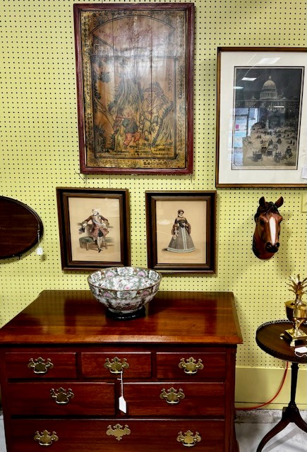 A vintage wooden chest of drawers is topped with a decorative floral bowl and a small golden figurine, perfect for those who appreciate antiques and collectables shopping. Hanging above are various framed artworks, including paintings and prints, alongside a mounted horse head sculpture on a pegboard wall.