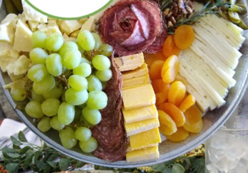 A platter filled with various cheeses, grapes, nuts, dried apricots, and slices of meat sits above a plate of thinly sliced roast beef topped with herbs and onions. The logo reads 