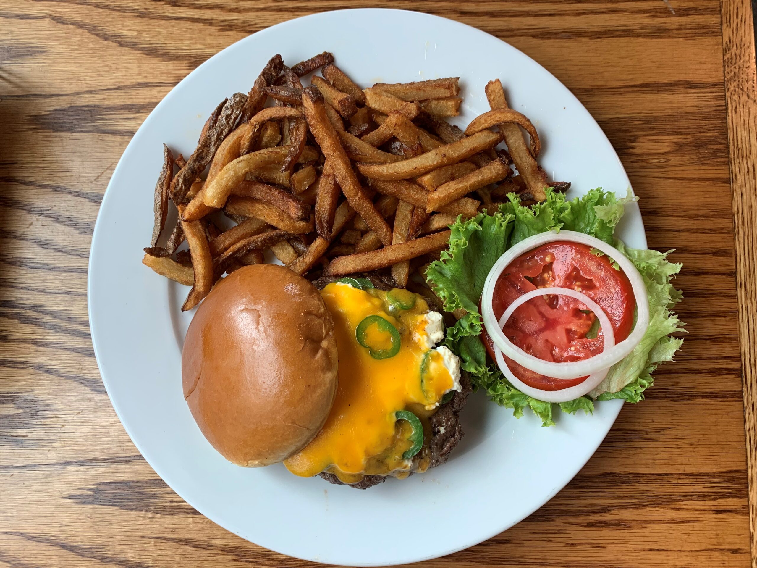A cheeseburger topped with melted cheese and jalapeños, served with a side of crispy french fries on a white plate. The burger includes fresh lettuce, tomato slices, and onion rings on the side—perfect for savoring during your weekend getaway. The plate rests on a wooden table.