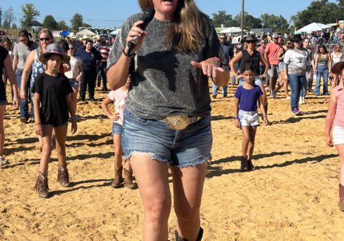 A woman in sunglasses, a gray t-shirt, denim shorts, and cowboy boots holds a microphone while walking on a sandy area. People, including children, are gathered around, with some wearing cowboy hats. The scene appears to be outdoors on a sunny day. Image