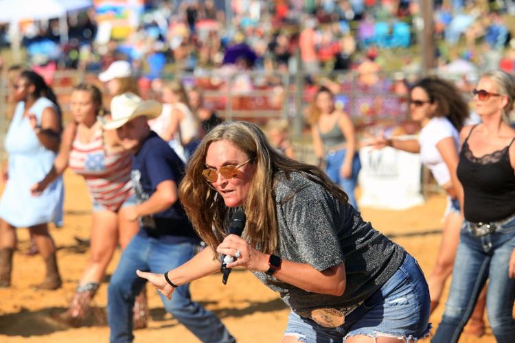 A woman holding a microphone leads a group of people in a dance at an outdoor event. She wears sunglasses, a grey shirt, and denim shorts. The participants, some in cowboy hats and Western attire, dance on a sandy surface with a large crowd watching in the background.