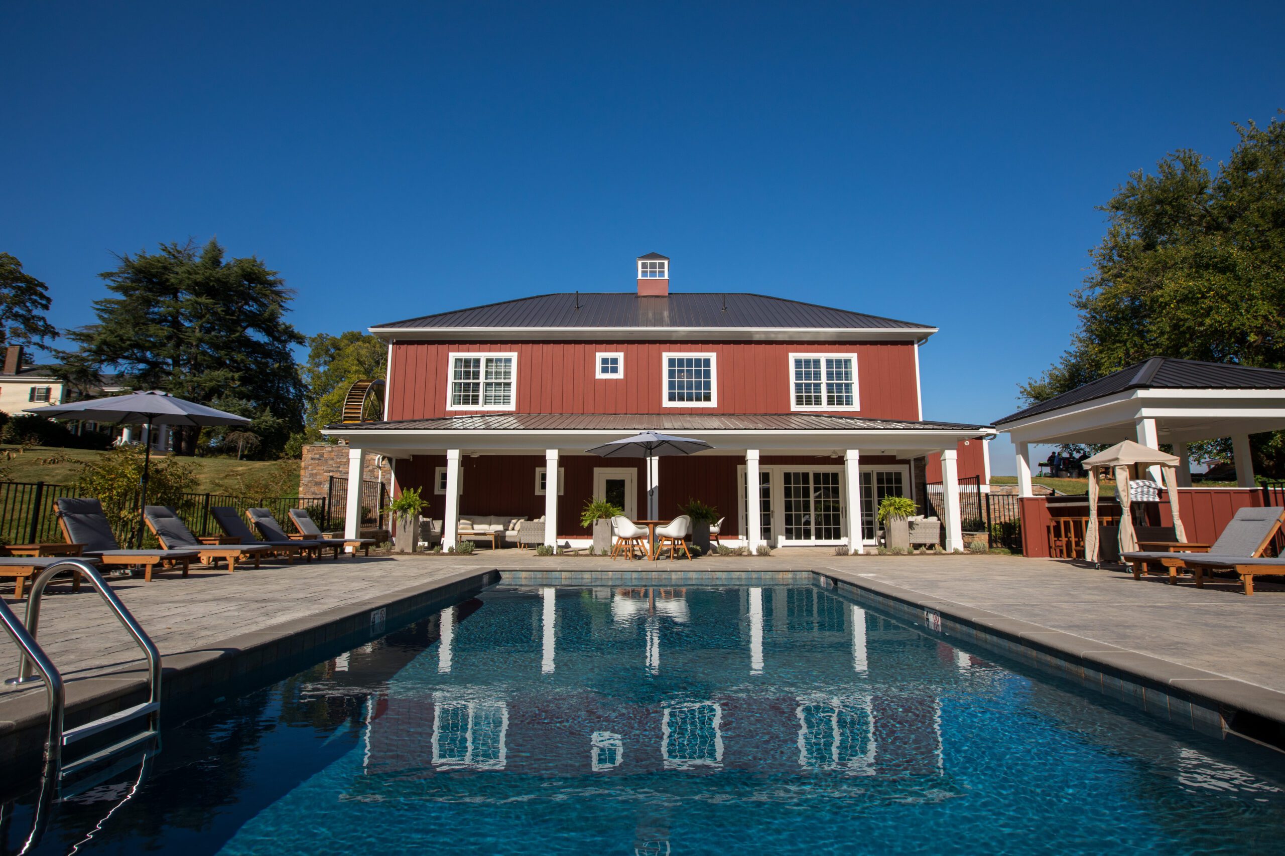 A large red barn-style building with a black roof stands under a clear blue sky. In front of it is a luxurious swimming pool with lounge chairs and umbrellas placed around it. The area is surrounded by greenery, creating a serene and inviting atmosphere.