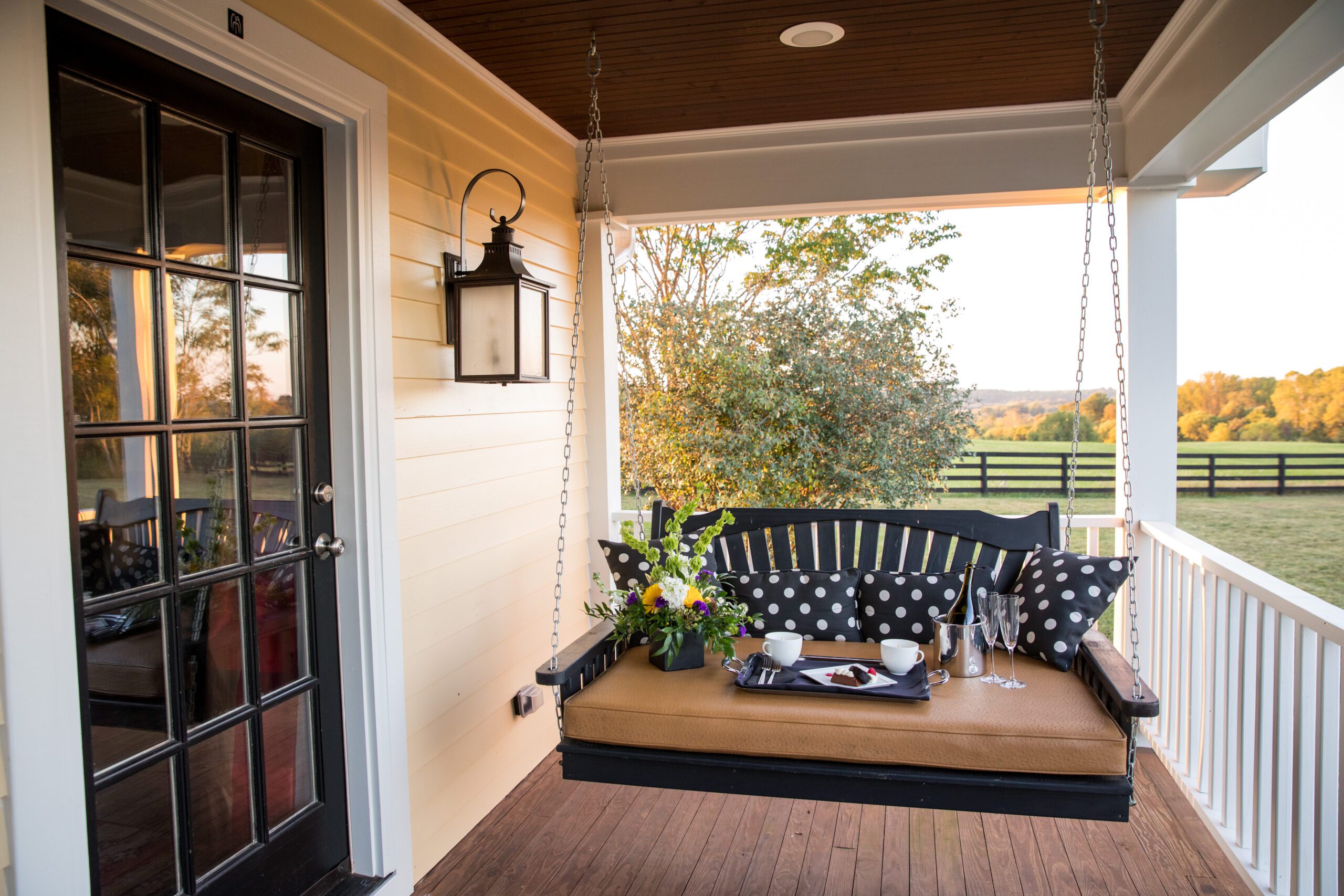 A porch with a cozy swing bench adorned with black and white polka-dot pillows and a tray holding flowers and teacups. A glass door is on the left, and the background shows a scenic view of trees and a meadow at sunset.