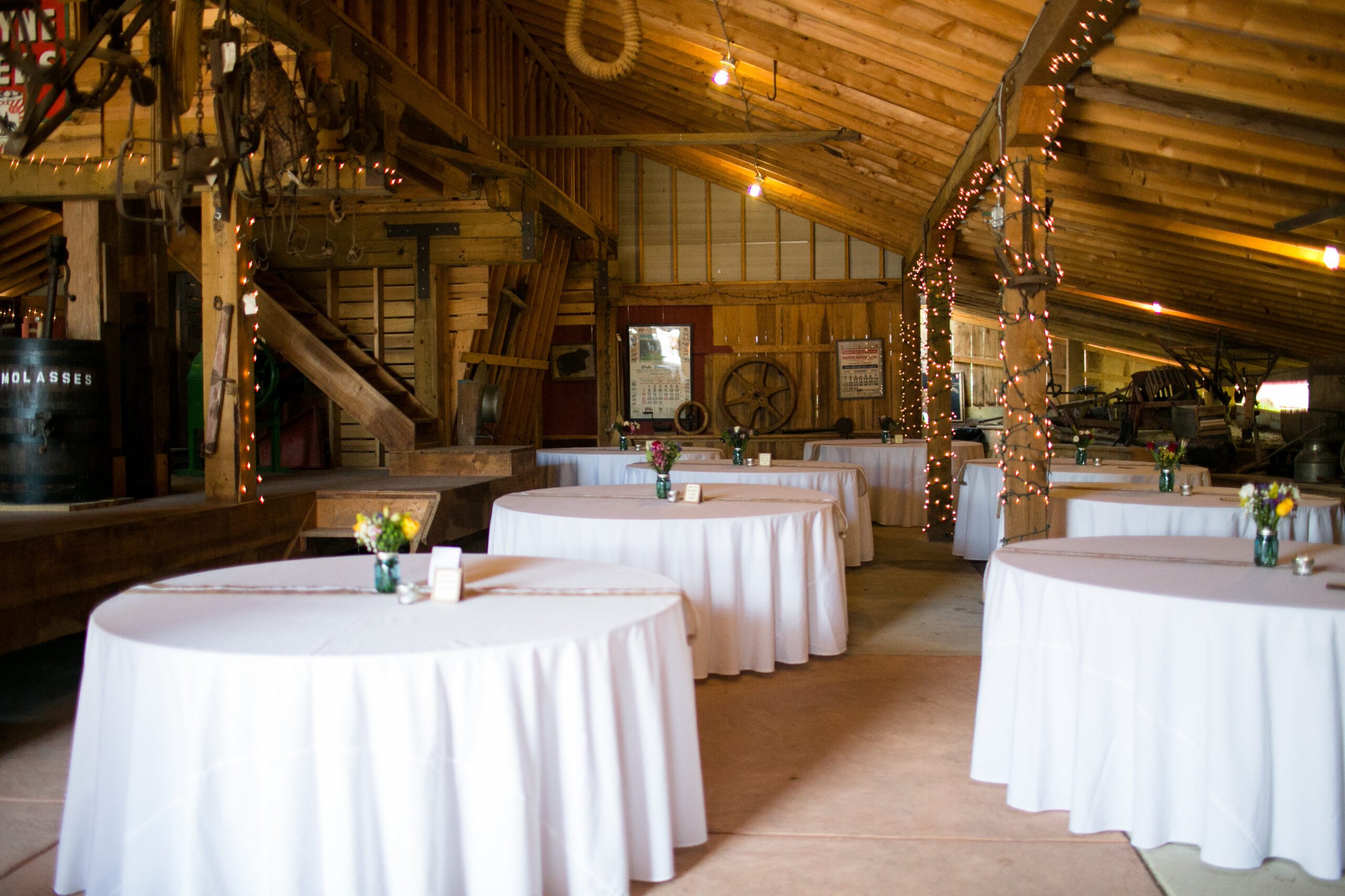 An indoor event venue with round tables covered in white tablecloths. The rustic wooden interior features string lights wrapped around central poles, and small flower arrangements are placed on each table. Various decorations and equipment are visible in the background, making it one of the charming wedding venues in Virginia.