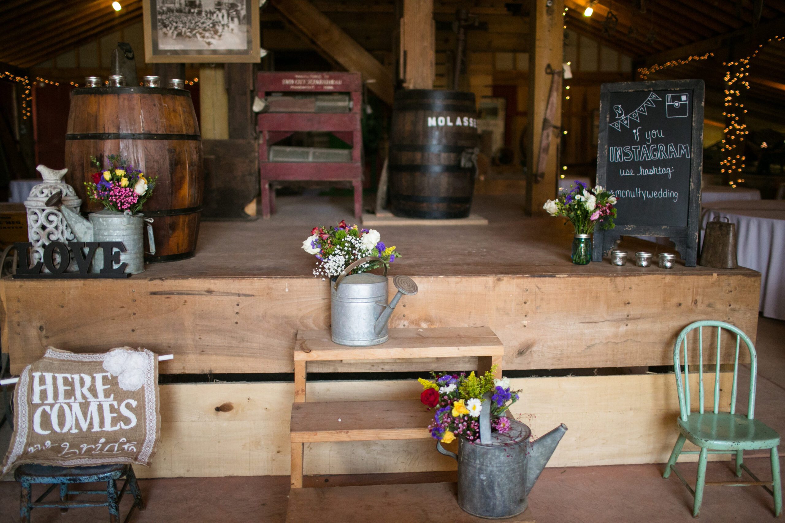 A rustic wedding setup featuring a wooden stage adorned with barrels and flowers, perfect for those seeking charming wedding venues in Virginia. A sign on the right reads “Instagram your hashtag #ourrusticwedding.” The front of the stage boasts vases and watering cans filled with flowers, while a pillow with 