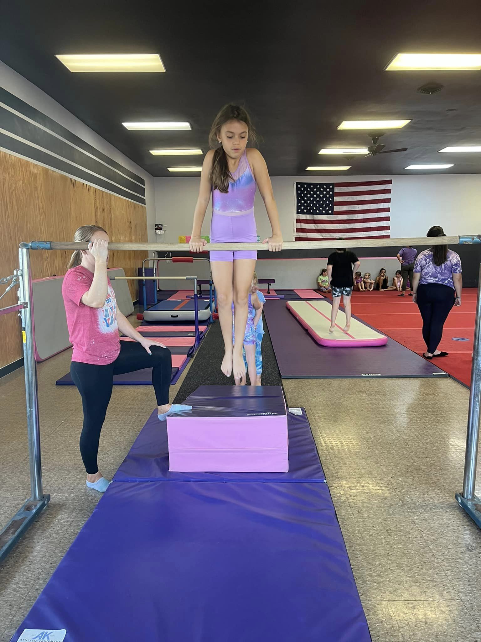 A young girl in a purple leotard practices on the uneven bars in a gymnastics facility. A coach in a red shirt and black pants assists her. Other children and trainers are visible in the background, with a large American flag hanging on the wall.