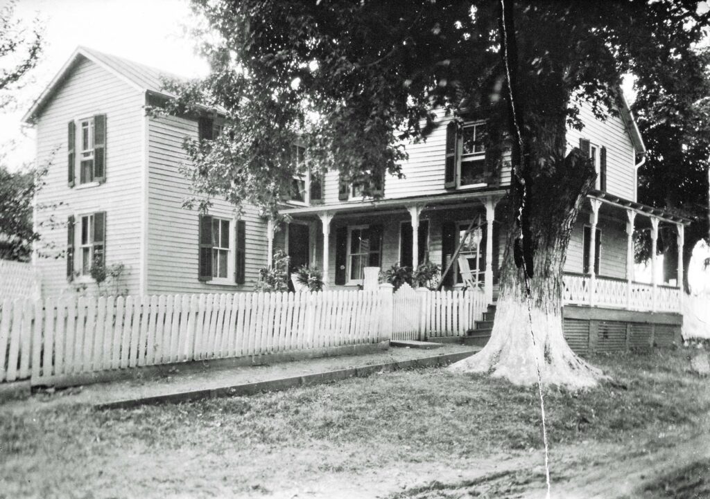 Black and white image of a two-story house with wooden siding and shutters. The house has a large porch with decorative trim, surrounded by a white picket fence. A large tree stands in the front yard. The overall scene appears to be from an earlier time period.