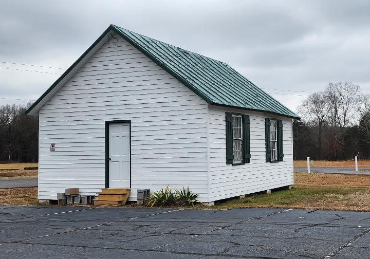 A small white wooden building with a green metal roof and green-framed windows. It sits on a gray, cracked pavement surrounded by grass. The sky is overcast, and bare trees are visible in the background. There are small steps leading to the front door.