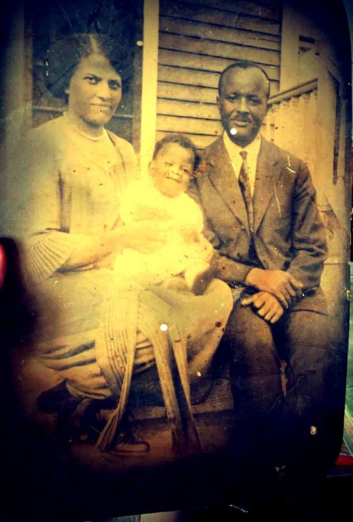 A sepia-toned vintage photograph shows a family of three sitting on steps in front of a wooden house. A woman on the left holds a baby, while a man in a suit sits to the right. All three are facing the camera and smiling.