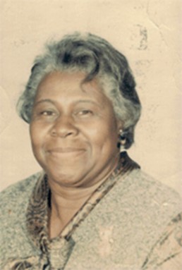 A portrait of an elderly woman with short, curly grey hair and a kind smile. She is wearing a long-sleeved, patterned blouse with a scarf tied around her neck. The background is plain and neutral-colored.