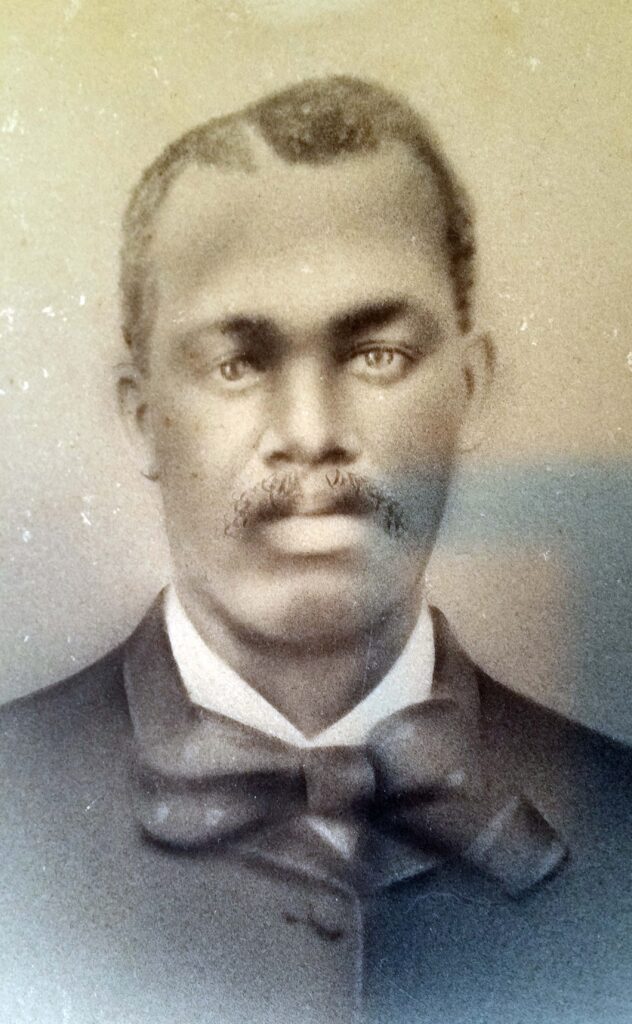 A sepia-toned portrait of a man with short hair and a mustache. He is dressed in a dark suit jacket, a white shirt, and a large dark bow tie. The image appears to have an old-fashioned, vintage quality.