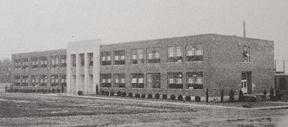 A black-and-white photo of a two-story brick building with multiple windows. The building has a central entrance characterized by a taller section with columns. The surrounding area appears to be an open field with some shrubbery along the building's facade.