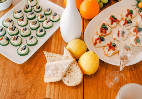 A wooden table set with an assortment of finger foods including cucumber bites on a white platter, lemon slices, bread, citrus fruits, and bruschetta. A white vase with a single flower and elegant glassware and plates complete the setup. Image