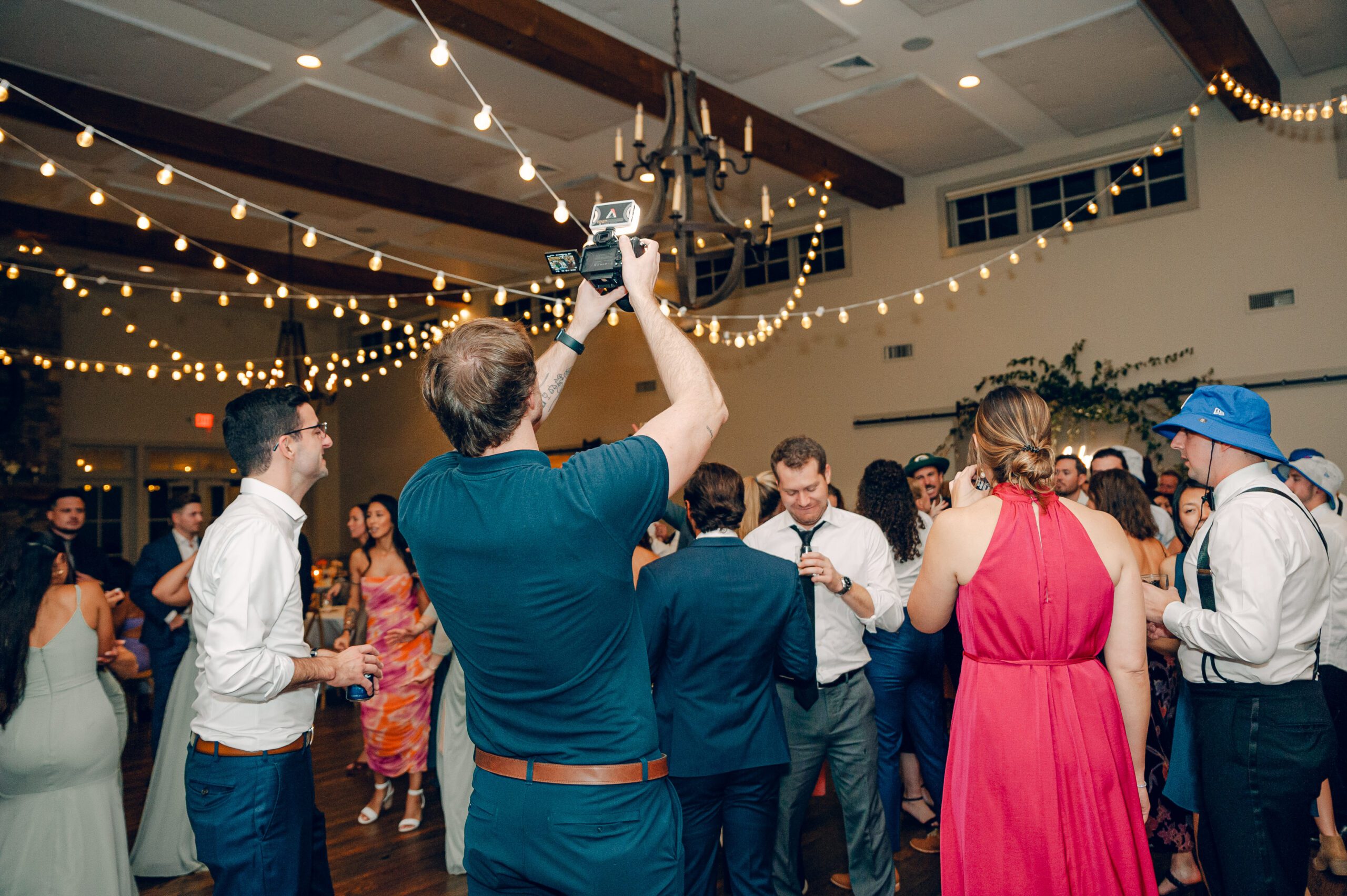A man holding a camera captures a lively moment on a dance floor filled with people dressed in formal attire. String lights hang from the ceiling, creating a festive atmosphere in a spacious room with wooden beams and chandeliers.