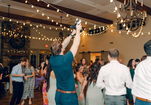 A lively indoor wedding reception with guests dancing on a wooden floor. One man is standing and holding a camera up to capture the moment. The room is decorated with string lights and chandeliers, and people are dressed in formal attire. Image